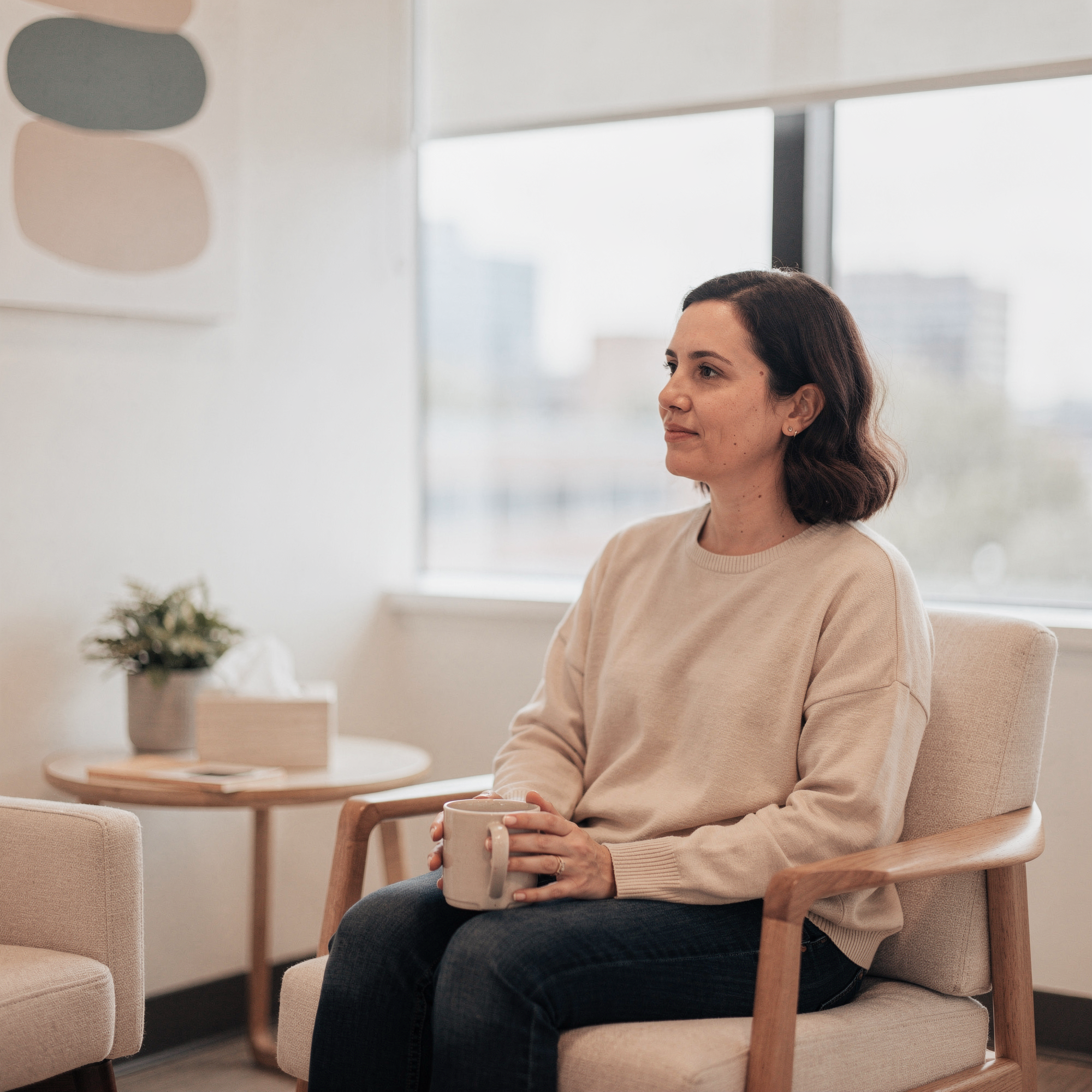 Woman sitting in a modern office lounge holding a mug, with city skyline visible through floor-to-ceiling windows